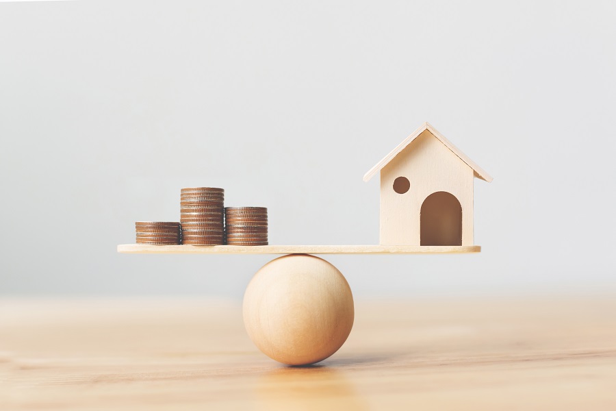 Wooden home and stack of coin resting on a scale, indicating the sale-demand seesaw.